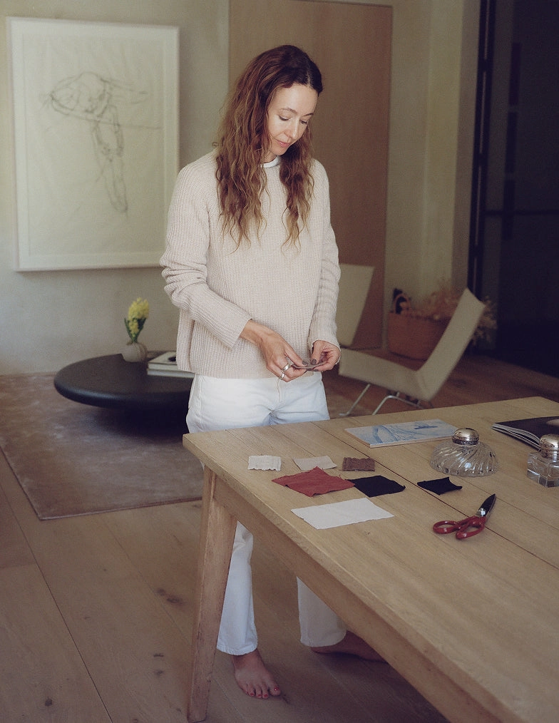 A woman in a cream sweater and white trousers leaning over a wood table with small objects