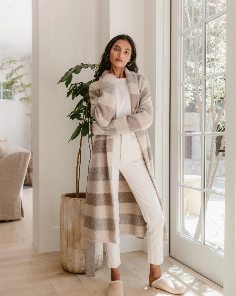 A woman in a long belted sweater coat and cropped jeans standing by a tall window next to a potted tree
