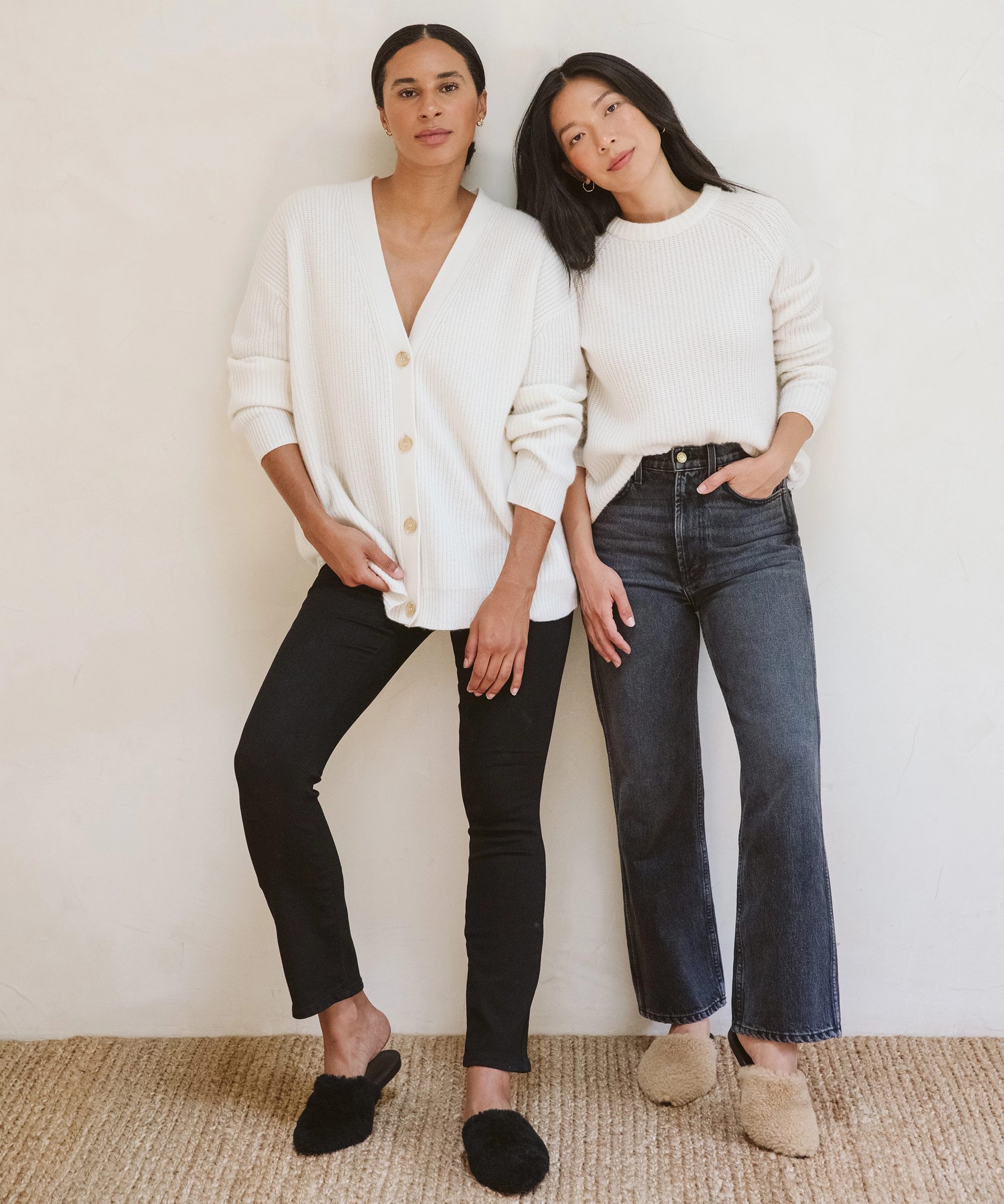 Two models in white knits and dark trousers standing next to each other against a white wall