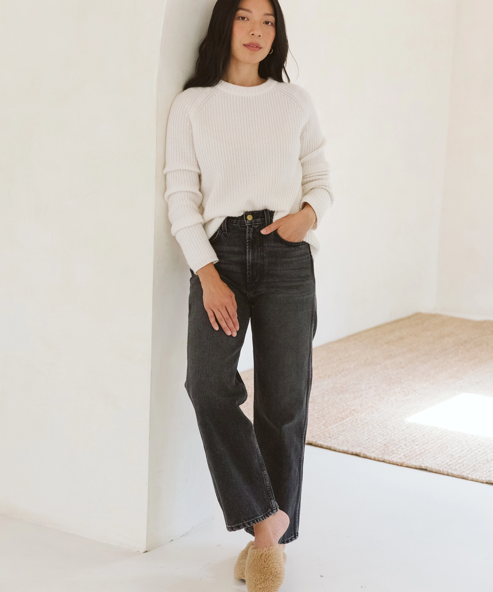 A woman with long dark hair leans against a white wall in a white ribbed sweater, high-waisted black jeans, and tan fuzzy Jenni Kayne mules
