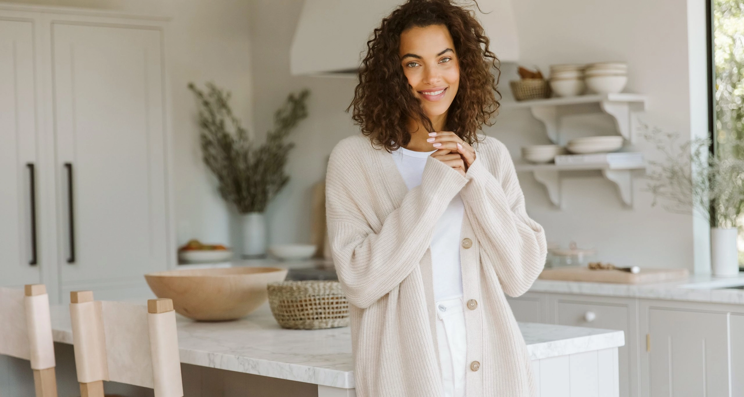 A smiling woman with curly hair in a light cardigan and white outfit stands in a bright modern kitchen with neutral decor