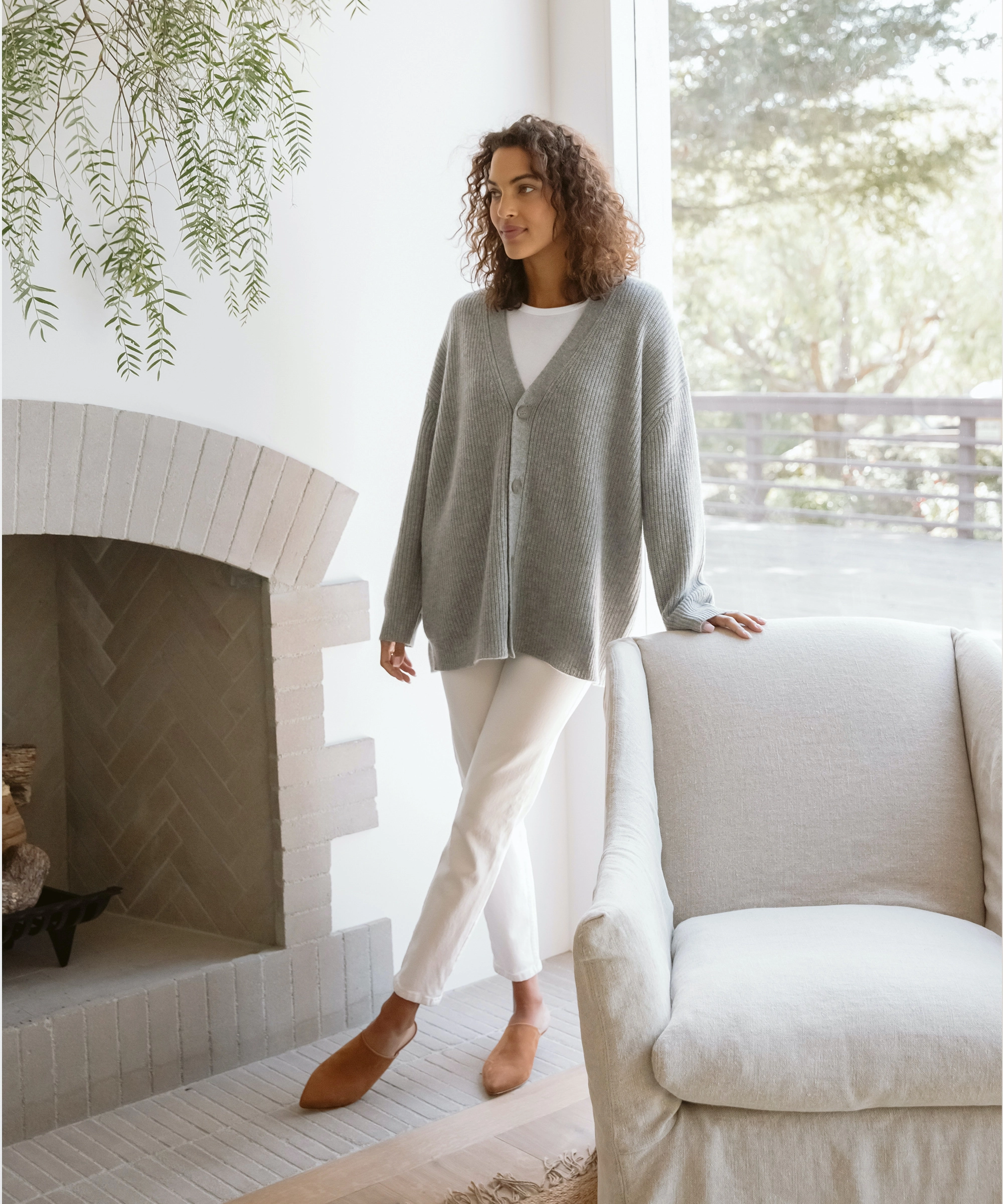A woman with curly hair stands by a large window in a sunlit living room in a gray cardigan, white pants, and brown shoes, one hand on a light armchair near the fireplace