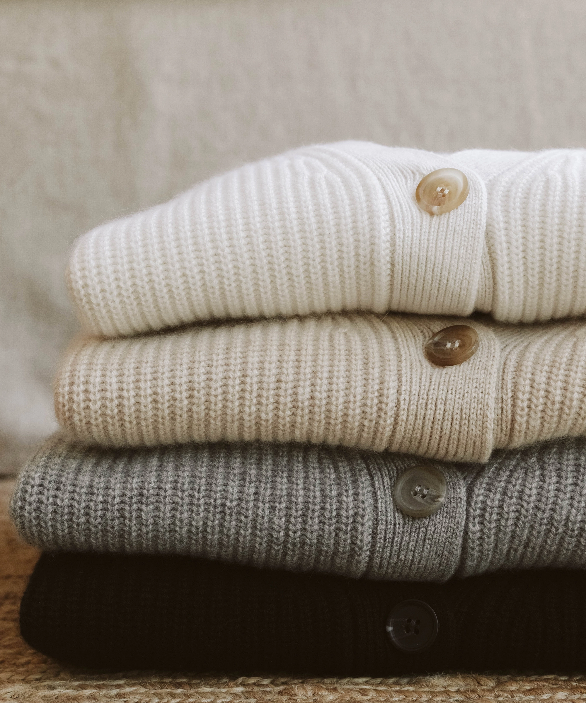 Four ribbed knit sweaters in white, beige, gray, and black folded in a stack, each with a visible button on top