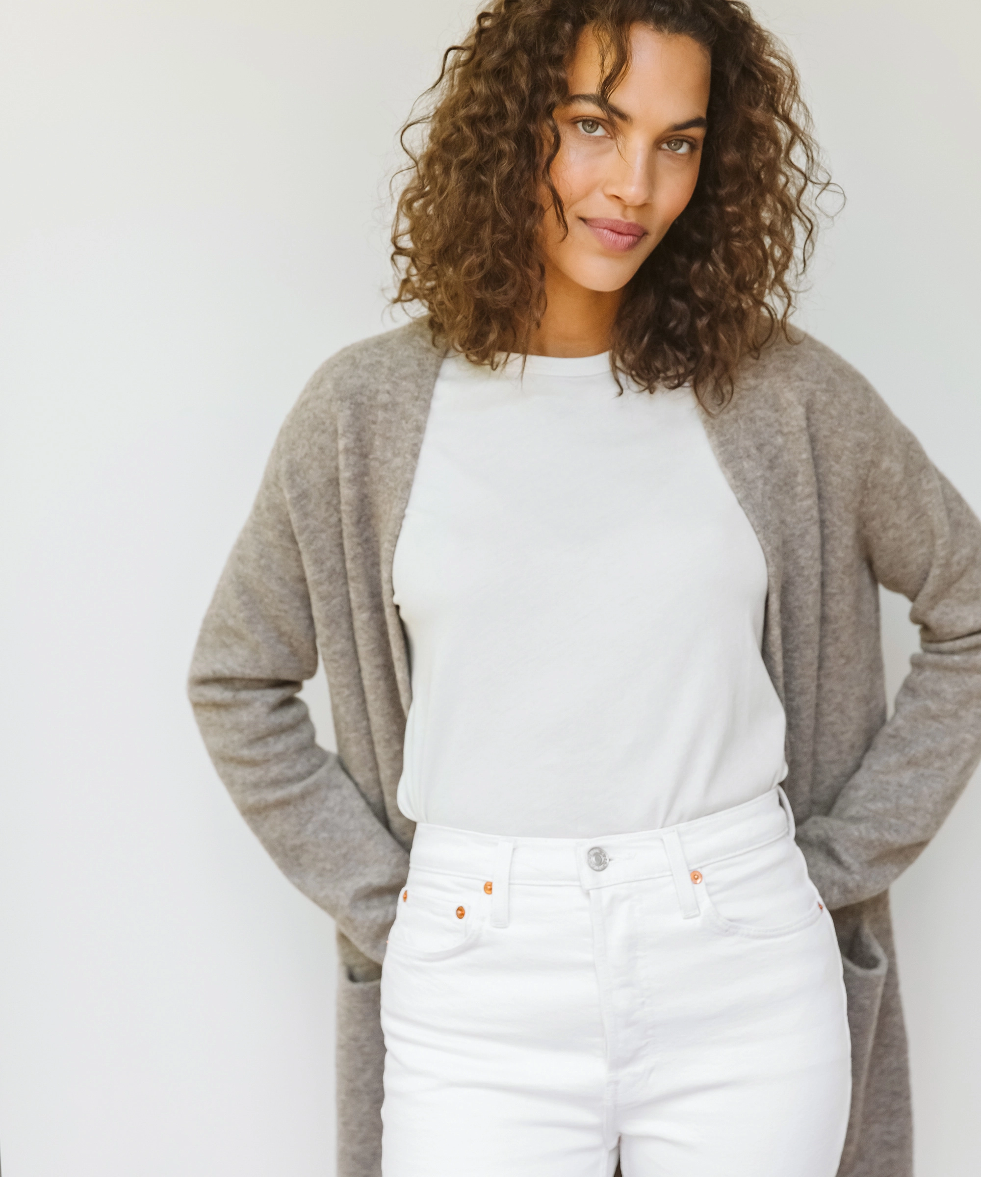 A woman with curly brown hair in a light gray cardigan over a white shirt and white jeans, smiling softly