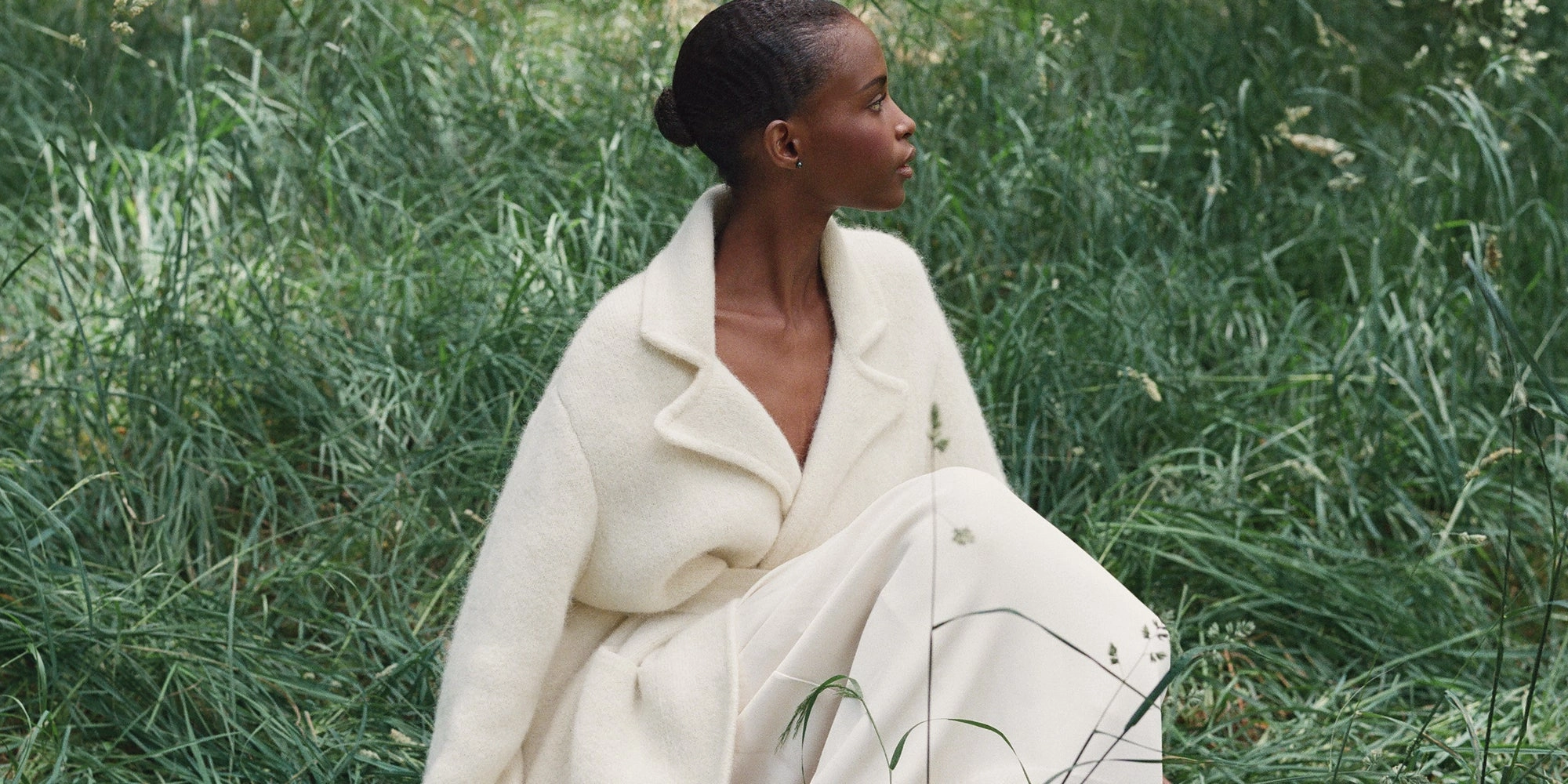 A woman in a cream wool coat sits in tall green grass and looks away from the camera