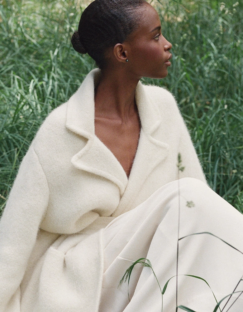 A woman in a cream wool coat seated in tall grass, looking to the side