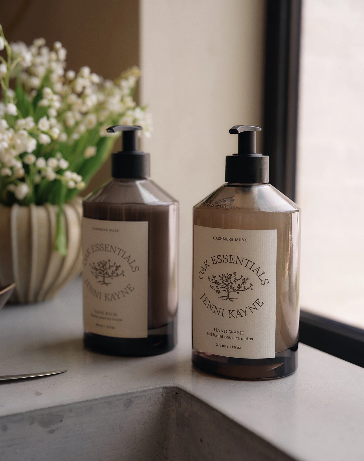 Two glass bottles of hand soap and lotion on a stone counter beside a vase of lily of the valley