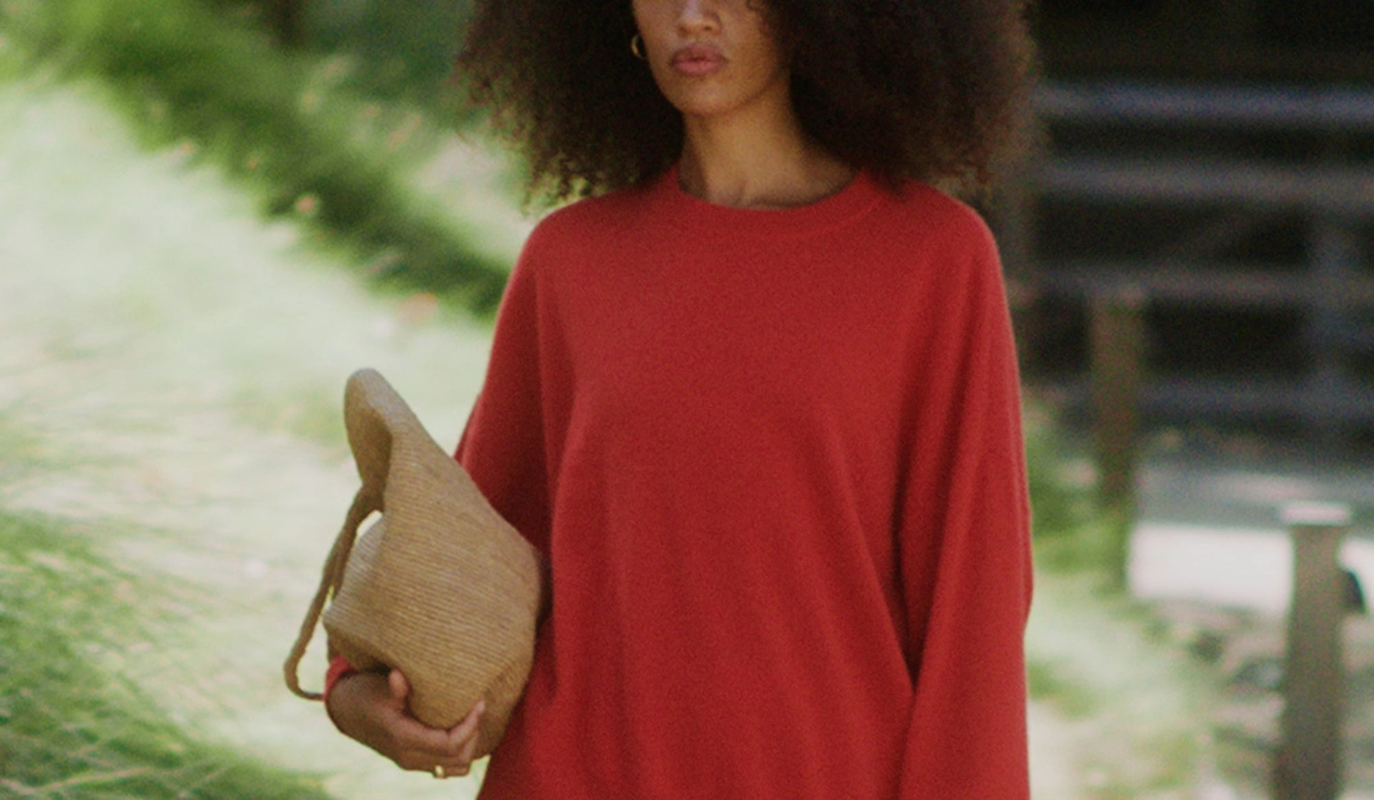 A woman in a red sweater holding a woven clutch, walking through soft green light